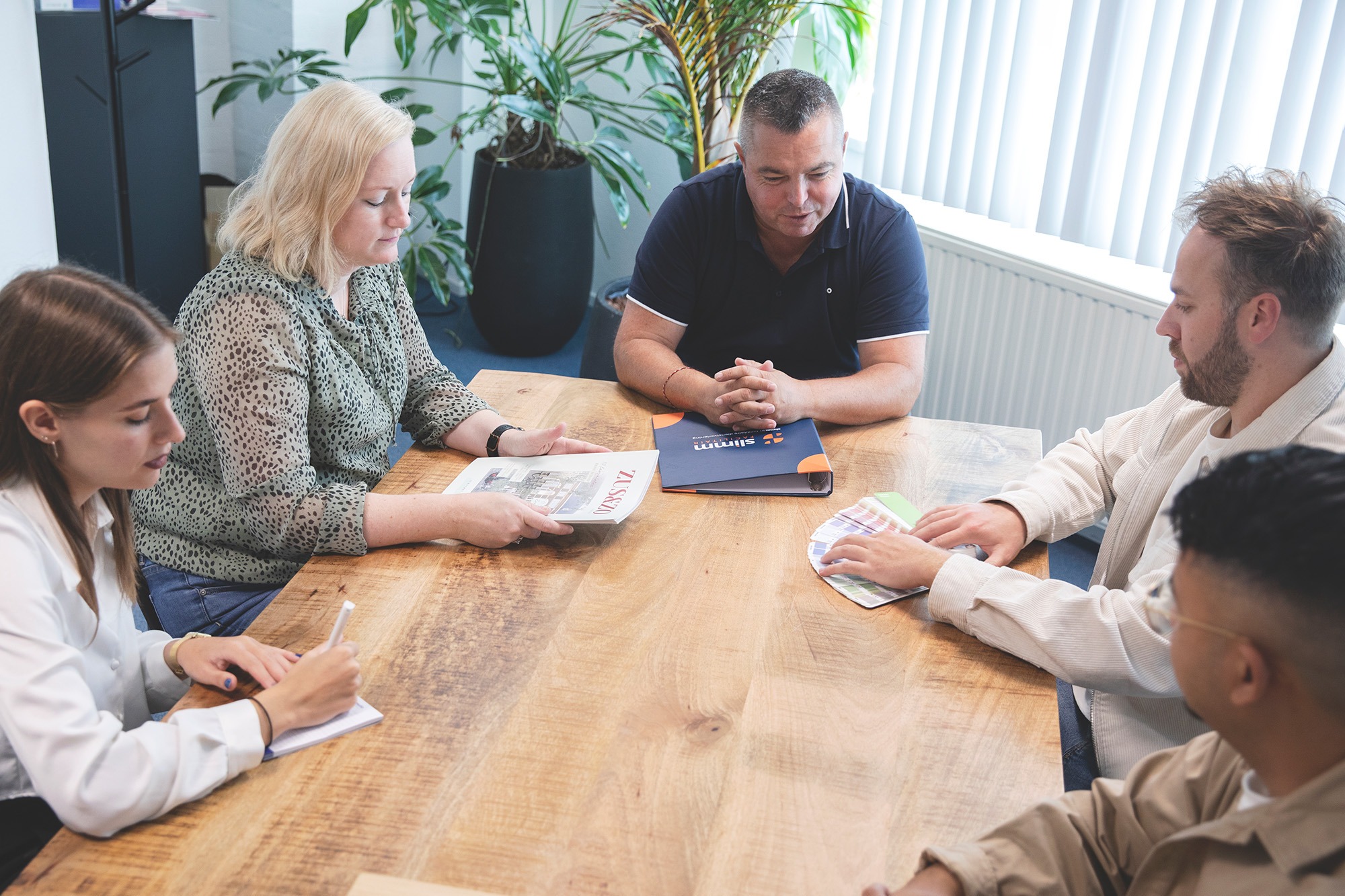 Team van Marker Media bespreekt een project met behulp van kleurstalen en drukwerk tijdens een creatieve vergadering aan tafel.